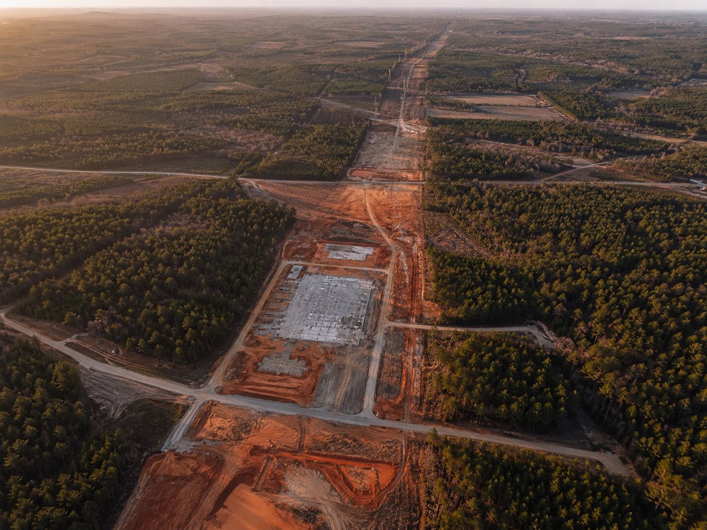 Aerial view of a rectangular concrete pad cleared from pine forest for a data center under construction, with a utility corridor cut through the trees.