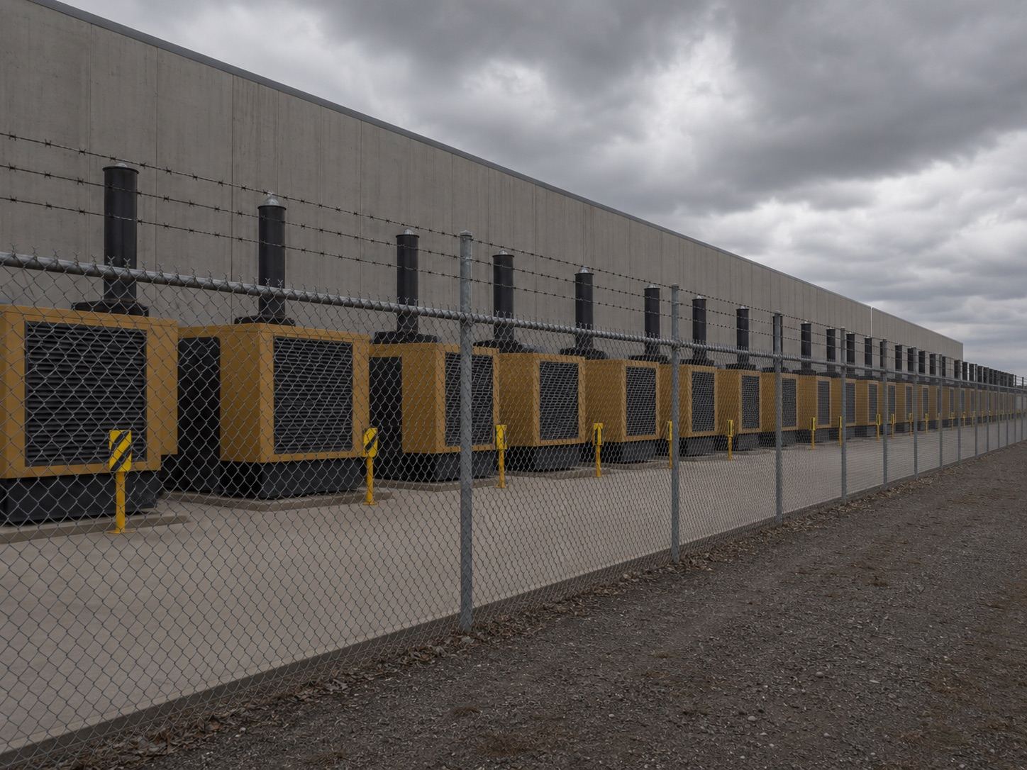 A long row of diesel backup generators behind a chain-link and barbed-wire fence along a data center building.