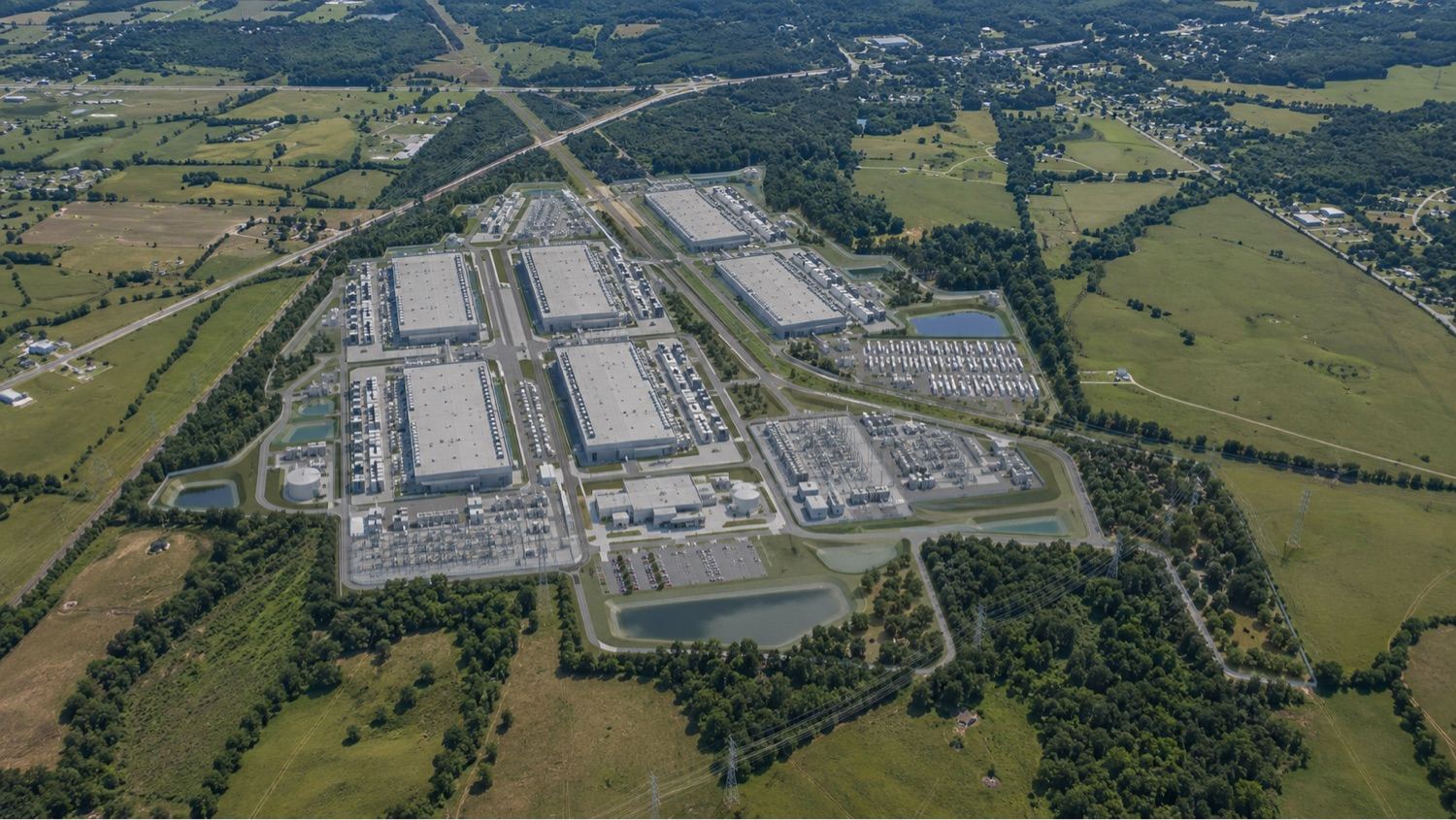 A photorealistic rendering of a hyperscale data center campus: several large flat-roofed buildings, a substation, retention ponds, and parking, set among farmland at a highway junction.
