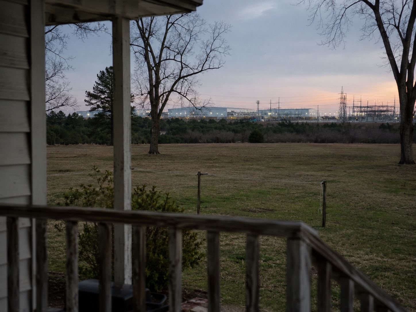 A view from a rural home's porch across a field toward a brightly lit data center campus and transmission towers at dusk.