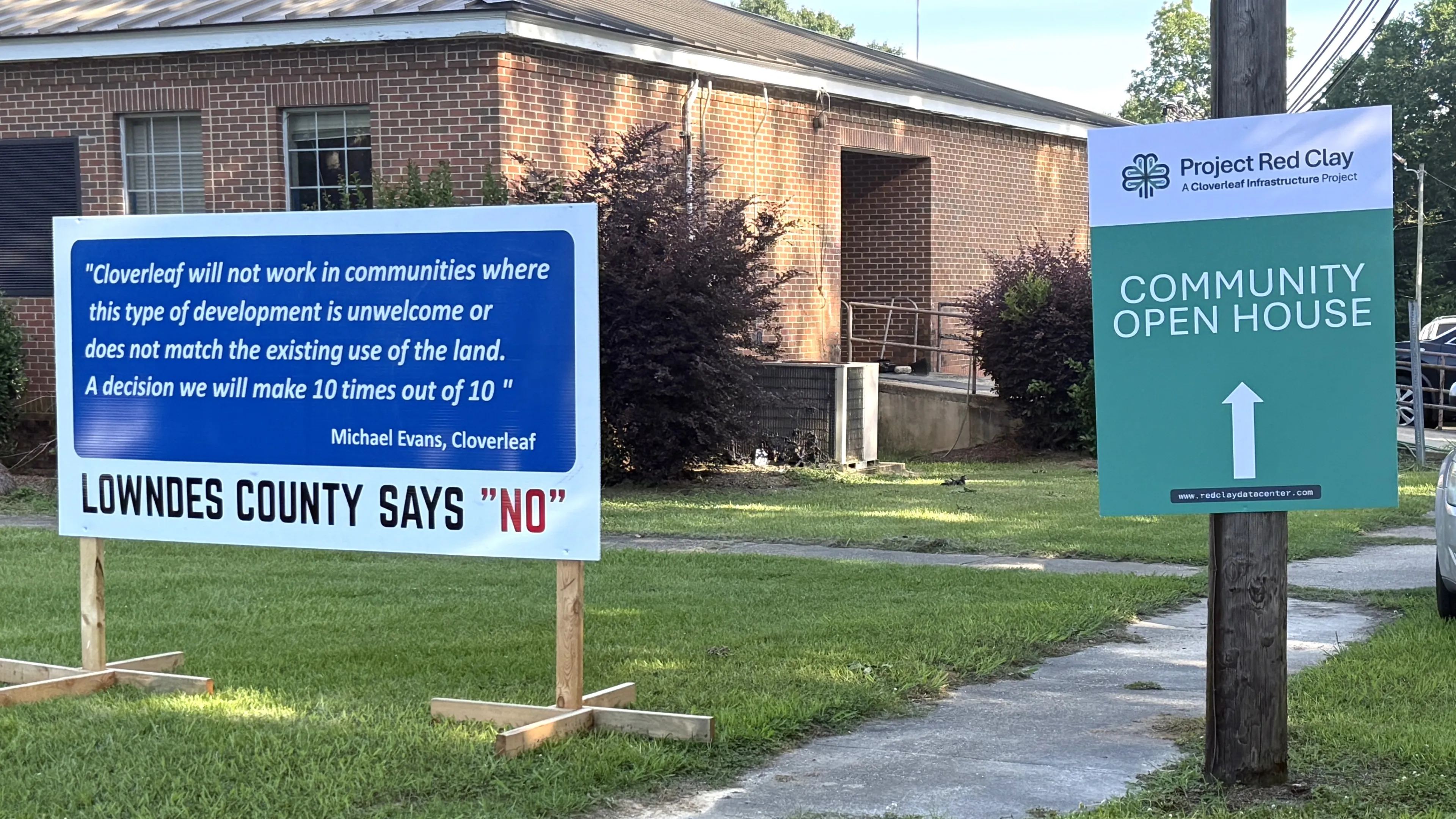 Two yard signs outside a brick building: one reading LOWNDES COUNTY SAYS "NO" over a quoted statement from a Cloverleaf representative, the other a green Project Red Clay sign directing visitors to a community open house.